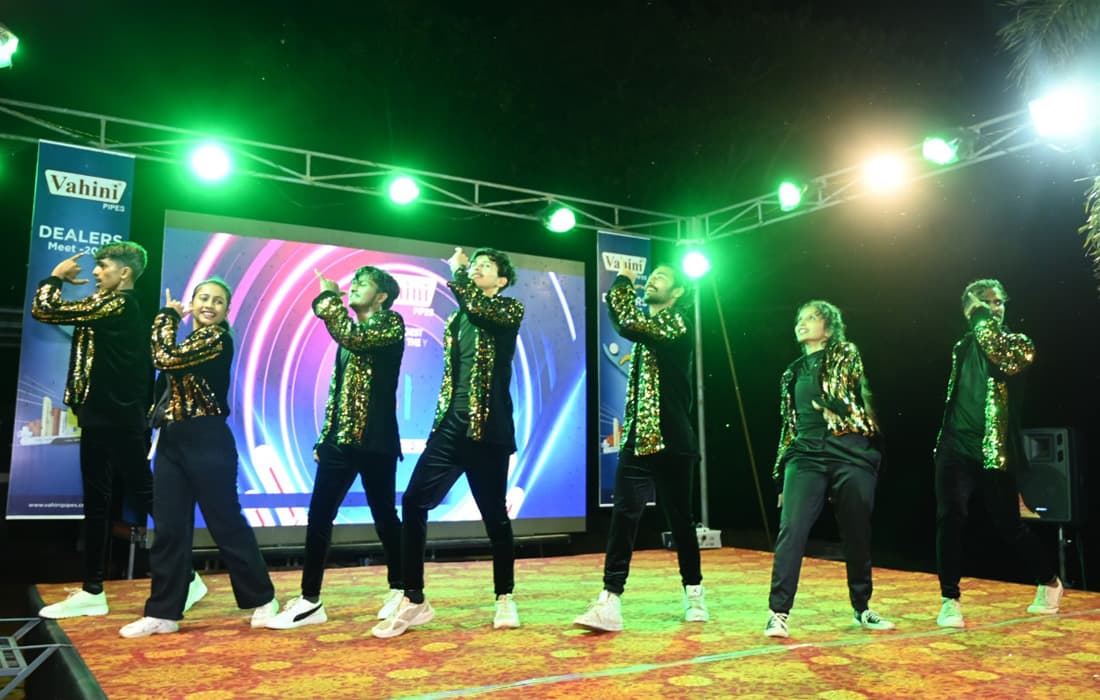 A dance troupe in sequinned jackets performing on an outdoor stage beside a Vahini Dealers Meet banner.