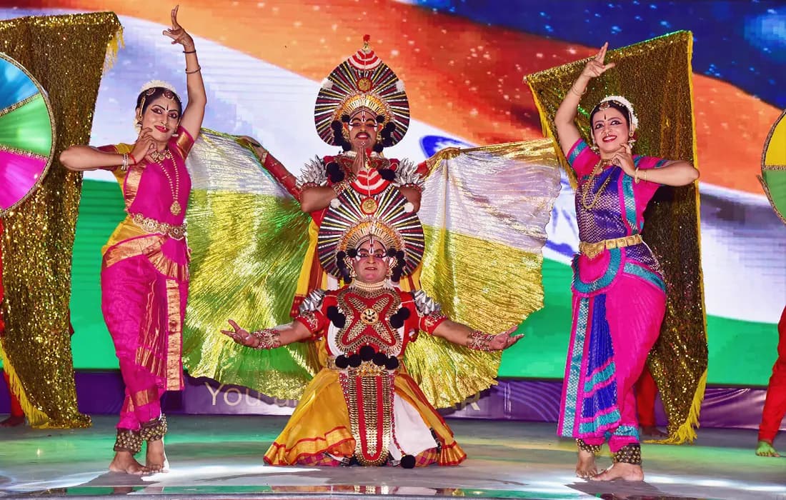 Classical and Yakshagana dancers performing on stage in front of the Indian flag.
