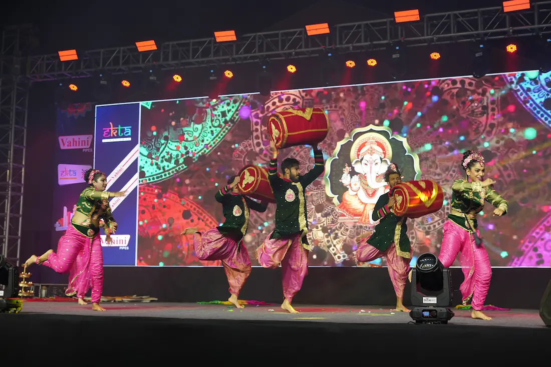 Dancers with drums performing on stage in front of a large screen showing a Ganesha graphic.