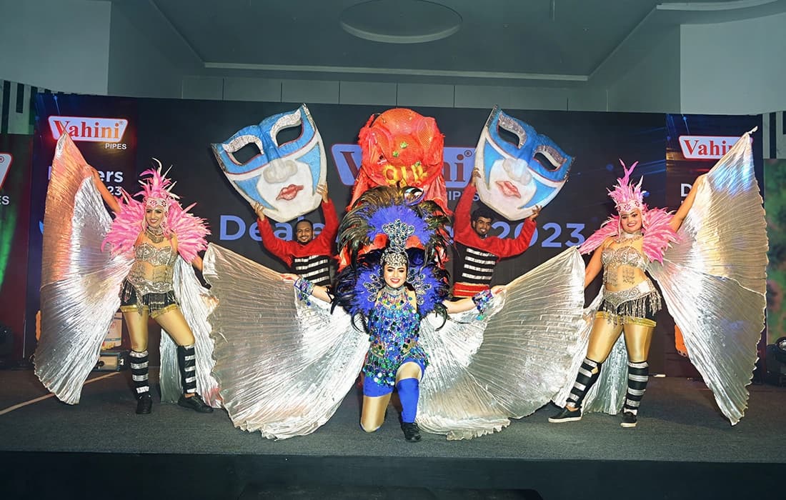 Stage performers in winged costumes and large masks in front of a Vahini Dealers Meet 2023 backdrop.