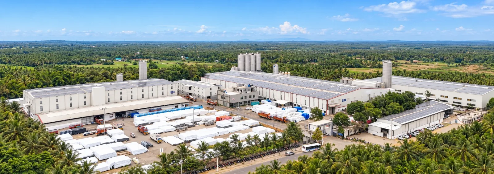An aerial view of the Vahini plant: long white production halls and silos, finished pipe and tanks stacked in the yard, coconut groves on every side.