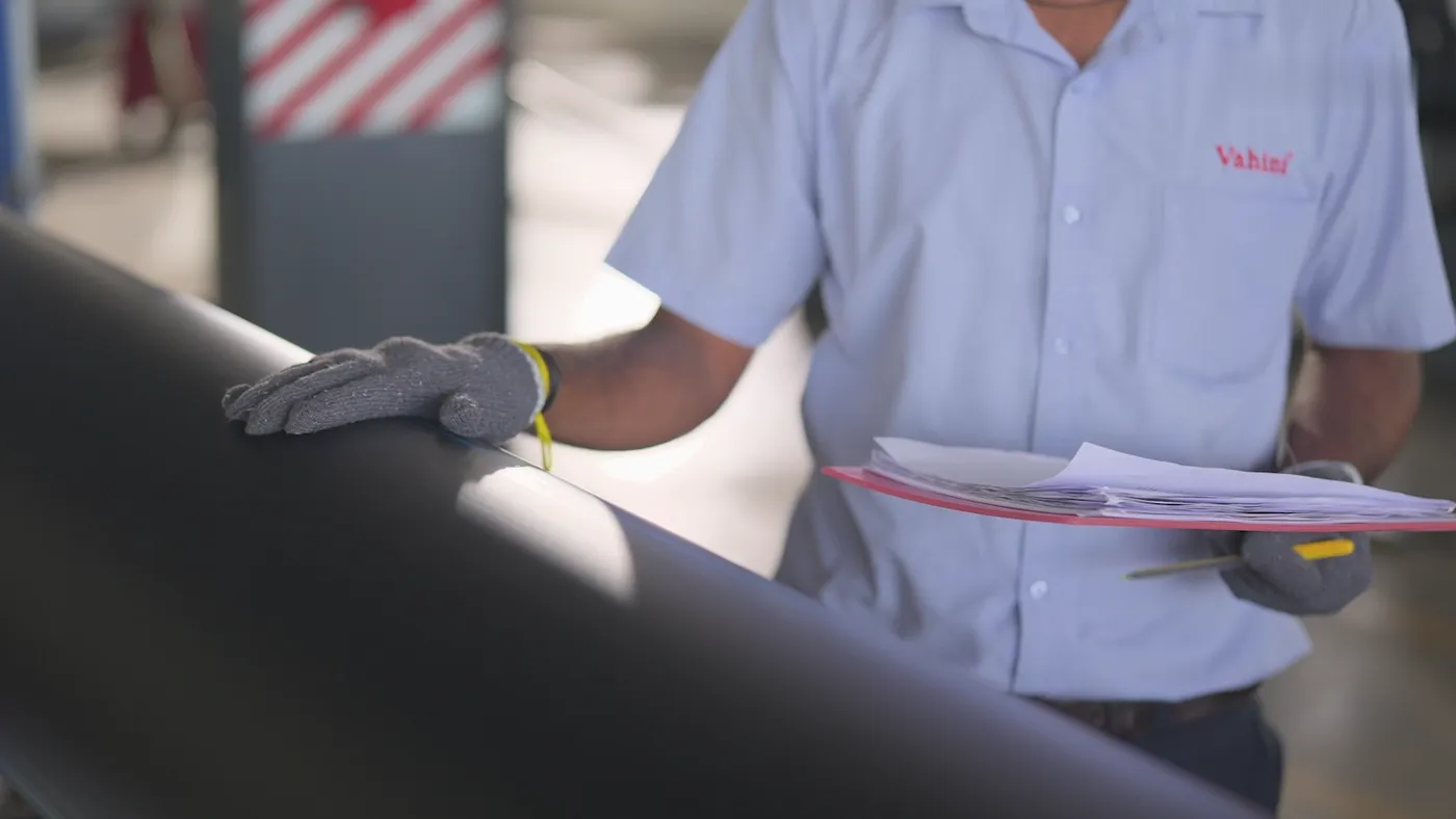 A member of staff in a Vahini shirt checking a length of black pipe, a clipboard of papers in one hand.